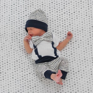 May include: A newborn outfit consisting of a white short-sleeved bodysuit with navy blue suspender straps, a matching navy blue and white dash-patterned bow tie, a matching beanie, and pants with navy blue cuffs and a white dash-patterned design.