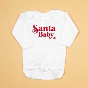 May include: White long-sleeved baby onesie with red text that reads "Santa Baby".