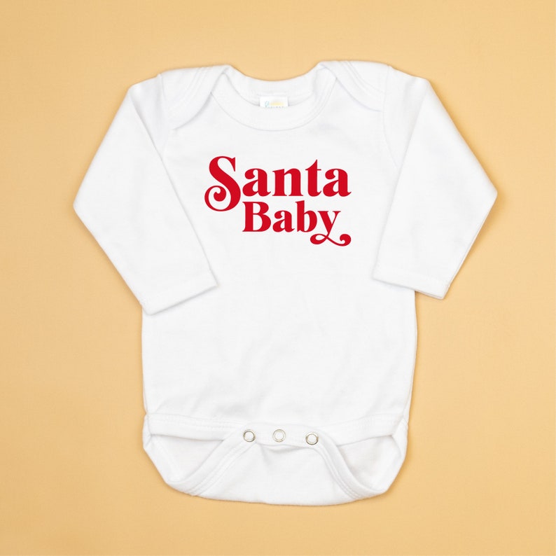 May include: White long-sleeved baby onesie with red text that reads "Santa Baby".