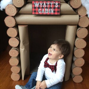 May include: A young boy wearing a white shirt with a red and black bow tie sits in front of a cardboard log cabin. The cabin is decorated with brown paper and has a sign that reads "Wyatt's Cabin" in white lettering on a red and black plaid background.