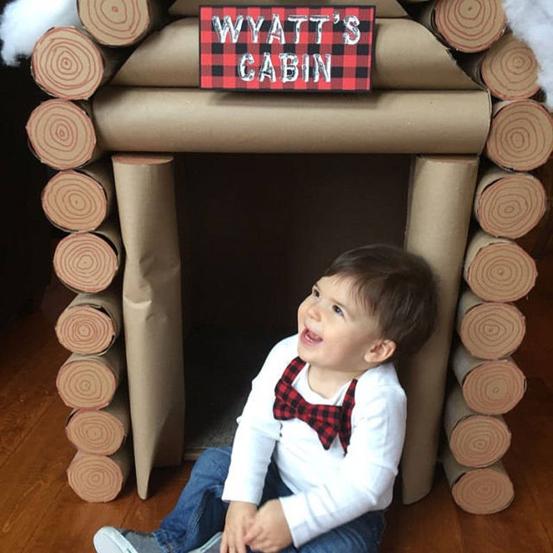 May include: A young boy wearing a white shirt with a red and black bow tie sits in front of a cardboard log cabin. The cabin is decorated with brown paper and has a sign that reads "Wyatt's Cabin" in white lettering on a red and black plaid background.