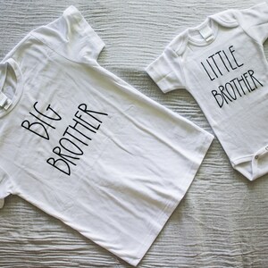 Op de afbeelding: Twee witte T-shirts met zwarte tekst. E&eacute;n shirt zegt "Grote Broer" en de andere zegt "Kleine Broer".