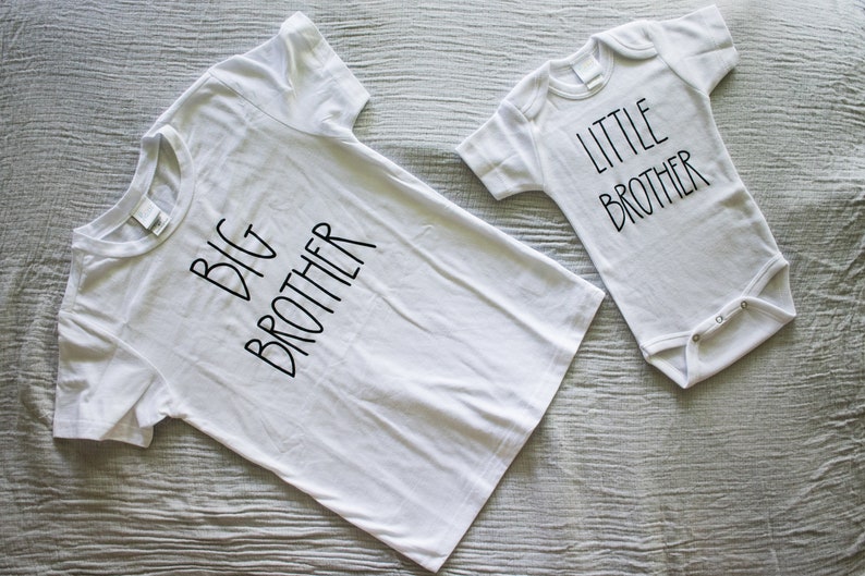Op de afbeelding: Twee witte T-shirts met zwarte tekst. E&eacute;n shirt zegt "Grote Broer" en de andere zegt "Kleine Broer".