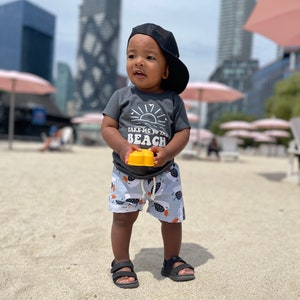 May include: A young child wearing a grey t-shirt with the text "Take me to the beach" and a black baseball cap. The child is holding a yellow toy and wearing blue and white patterned shorts.