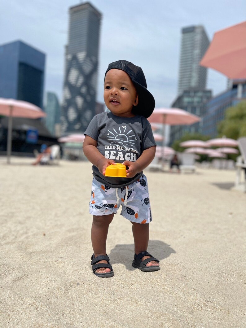 May include: A young child wearing a grey t-shirt with the text "Take me to the beach" and a black baseball cap. The child is holding a yellow toy and wearing blue and white patterned shorts.