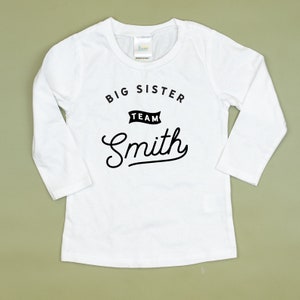 Puede incluir: Camiseta blanca de manga larga con texto negro que dice "Big Sister Team Smith".