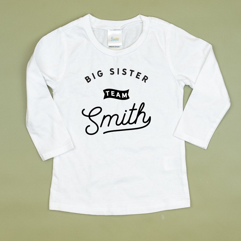 Puede incluir: Camiseta blanca de manga larga con texto negro que dice "Big Sister Team Smith".