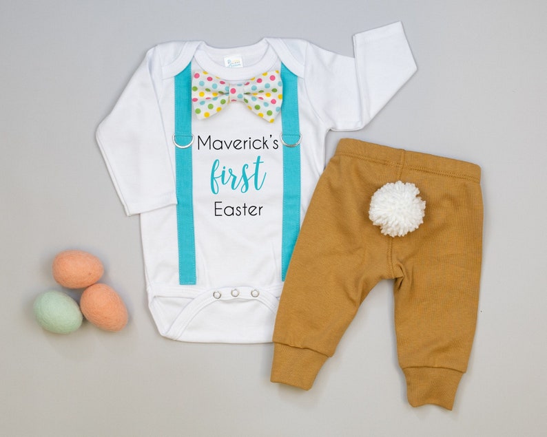 Puede incluir: Mono blanco para beb&eacute; con tirantes azules y una pajarita de lunares multicolores. El mono dice "Maverick's first Easter". Pantalones marrones con una cola de pomp&oacute;n blanco.