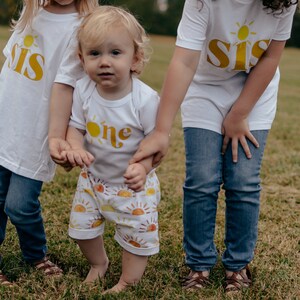 May include: Three children wearing white shirts with yellow sun and text designs. The youngest child is wearing a white shirt with the word "one" and yellow sun design. The two older children are wearing white shirts with the word "sis" and yellow sun design. The children are standing in a grassy field.