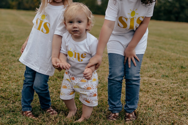 May include: Three children wearing white shirts with yellow sun and text designs. The youngest child is wearing a white shirt with the word "one" and yellow sun design. The two older children are wearing white shirts with the word "sis" and yellow sun design. The children are standing in a grassy field.