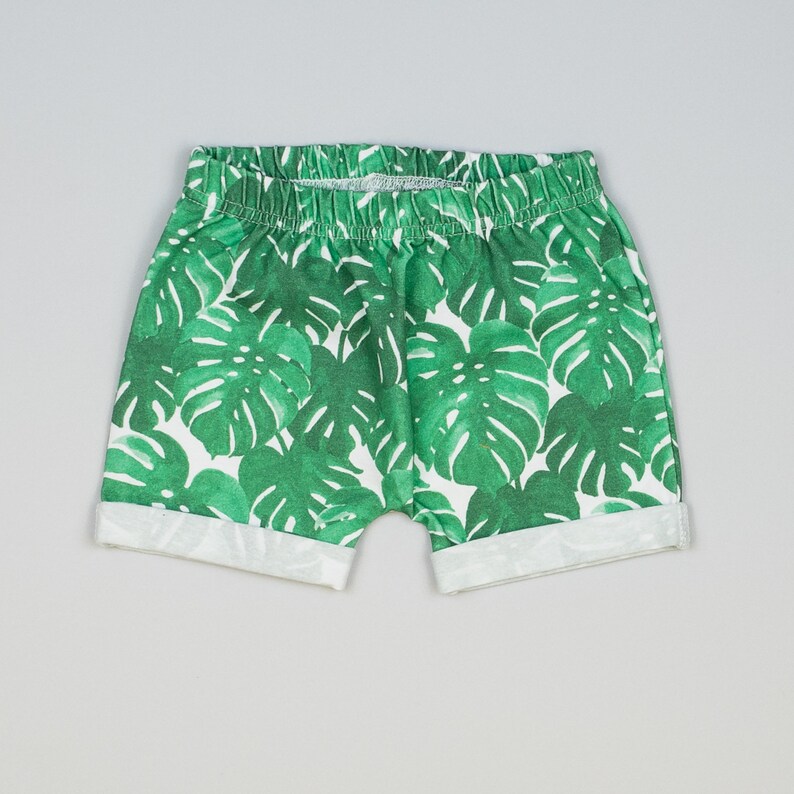 May include: White shorts with a green tropical leaf print. The shorts have a white elastic waistband and a cuffed hem.