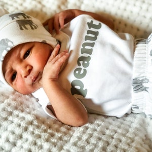 May include: A newborn baby wearing a white onesie with the word "peanut" printed on it in gray. The baby is also wearing a white and gray hat with a pattern of elephants. The baby is lying on a white blanket.