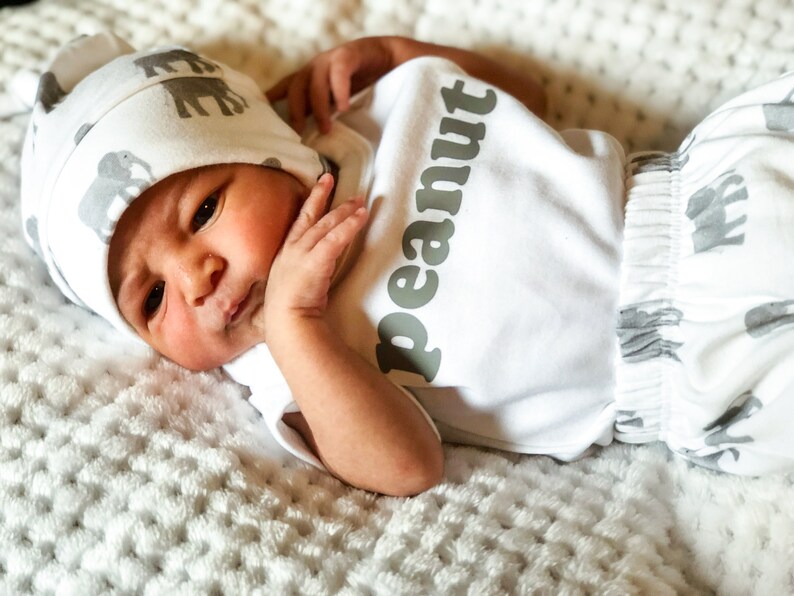 May include: A newborn baby wearing a white onesie with the word "peanut" printed on it in gray. The baby is also wearing a white and gray hat with a pattern of elephants. The baby is lying on a white blanket.