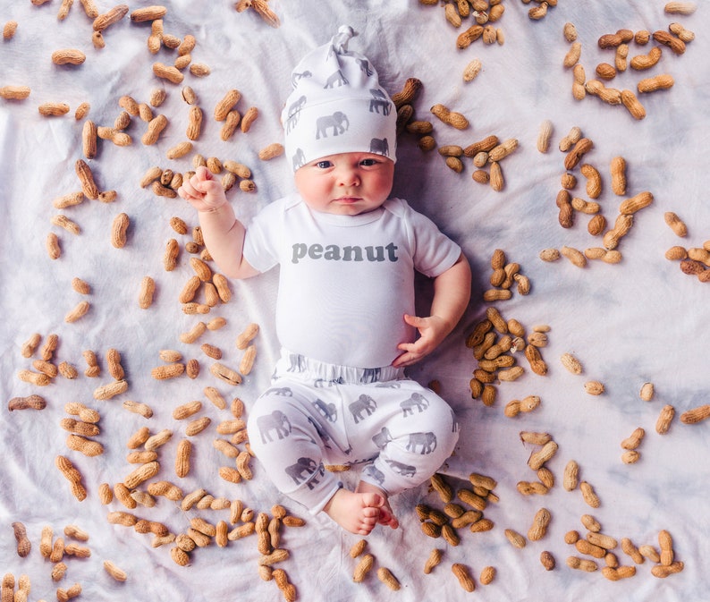 May include: A baby wearing a white onesie with the word "peanut" printed on it. The baby is wearing a gray and white hat with elephants printed on it. The baby is lying on a white blanket surrounded by peanuts.