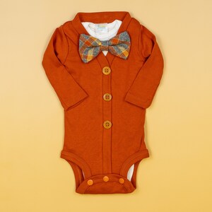 Op de afbeelding: Een roestkleurige, langarmige romper met een knoopsluiting en een geruite strik. De romper heeft een wit onderhemd en is gemaakt van een zachte, comfortabele stof.