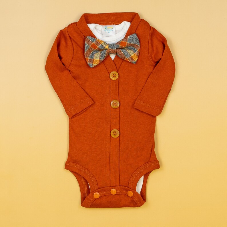 Op de afbeelding: Een roestkleurige, langarmige romper met een knoopsluiting en een geruite strik. De romper heeft een wit onderhemd en is gemaakt van een zachte, comfortabele stof.