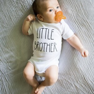 Op de afbeelding: Een baby draagt een wit rompertje met de tekst "LITTLE BROTHER" in zwarte letters. De baby ligt op een grijze deken en heeft een speen in de mond.
