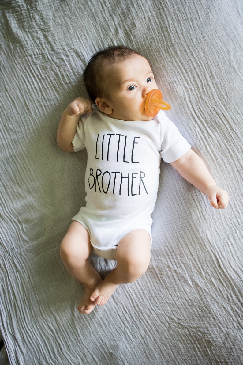 Op de afbeelding: Een baby draagt een wit rompertje met de tekst "LITTLE BROTHER" in zwarte letters. De baby ligt op een grijze deken en heeft een speen in de mond.