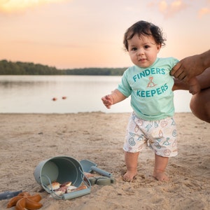 May include: A toddler wearing a light blue shirt with the text "Finders Keepers" and white shorts with a seashell pattern stands on a sandy beach. The child is holding a blue bucket and shovel.