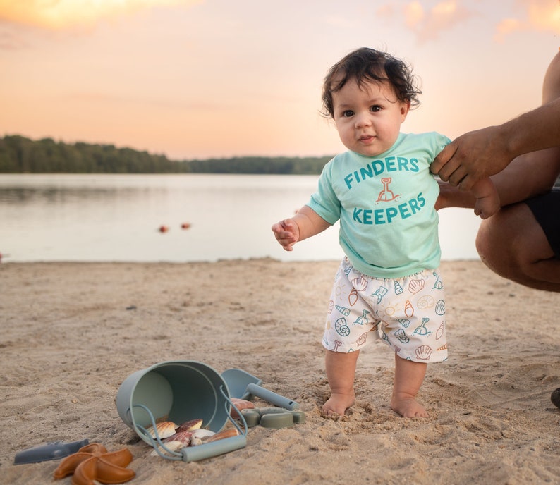 May include: A toddler wearing a light blue shirt with the text "Finders Keepers" and white shorts with a seashell pattern stands on a sandy beach. The child is holding a blue bucket and shovel.