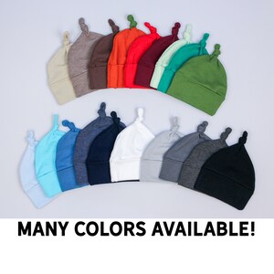May include: A selection of baby knot hats in a variety of colours, such as beige, brown, orange, red, teal, green, light blue, blue, navy, white, grey, dark grey, and black. The text "MANY COLORS AVAILABLE!" is displayed at the bottom.