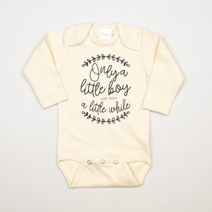 Pode incluir: Um macac&atilde;o de beb&ecirc; de manga comprida de cor creme com texto preto que diz "Only a little boy -for such- a little while" rodeado por uma coroa de folhas.
