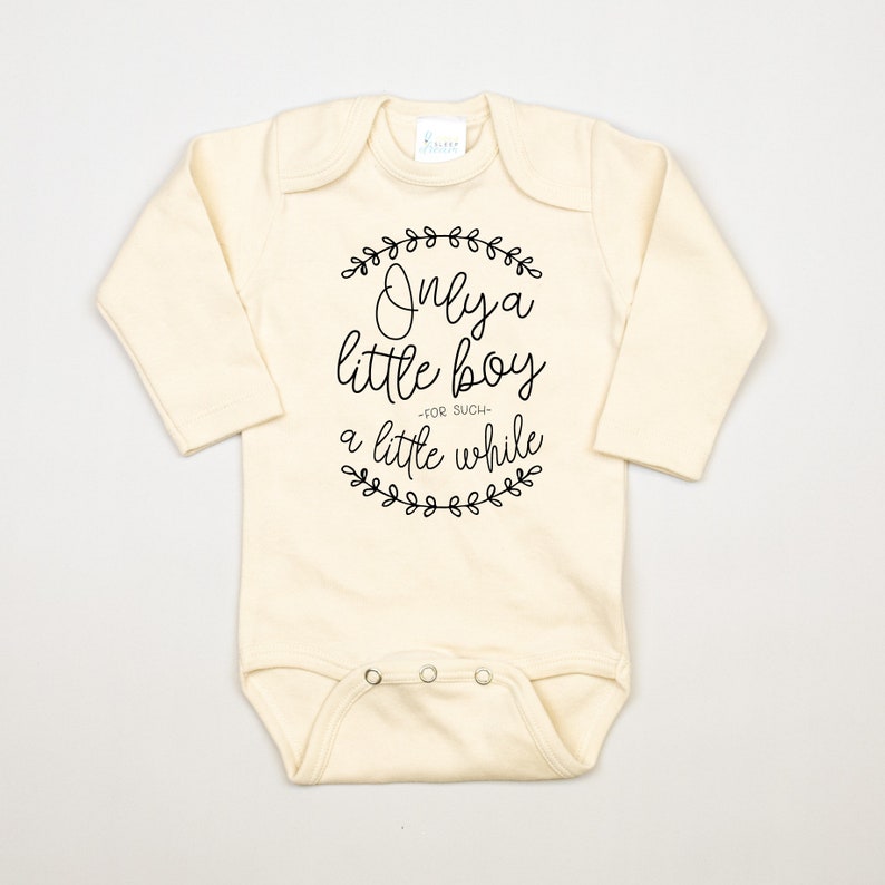 Pode incluir: Um macac&atilde;o de beb&ecirc; de manga comprida de cor creme com texto preto que diz "Only a little boy -for such- a little while" rodeado por uma coroa de folhas.