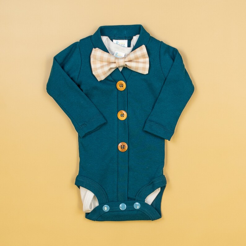 Op de afbeelding: Een baby-onesie in tealblauw met een lange mouwen cardigan-stijl top en een wit onderhemd. De onesie heeft een knoopsluiting aan de voorkant en een bruin geruite strikje.