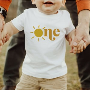May include: A white t-shirt with a yellow sun and the word "one" in a retro font.