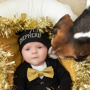Puede incluir: Un beb&eacute; con un gorro de punto negro con la palabra "SHEPHERD" bordada, una pajarita negra y un atuendo negro. El beb&eacute; est&aacute; acostado en una guirnalda amarilla y dorada con un perro marr&oacute;n y blanco olfateando la cara del beb&eacute;.