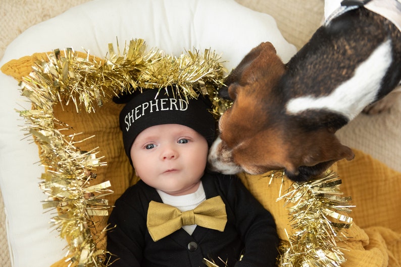 Puede incluir: Un beb&eacute; con un gorro de punto negro con la palabra "SHEPHERD" bordada, una pajarita negra y un atuendo negro. El beb&eacute; est&aacute; acostado en una guirnalda amarilla y dorada con un perro marr&oacute;n y blanco olfateando la cara del beb&eacute;.