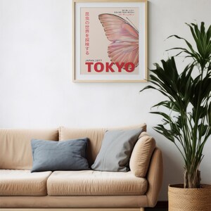 May include: A framed poster with a pink butterfly wing design and the text "JAPAN 1977 TOKYO" in red. The poster is hanging on a white wall above a beige couch with two gray pillows.