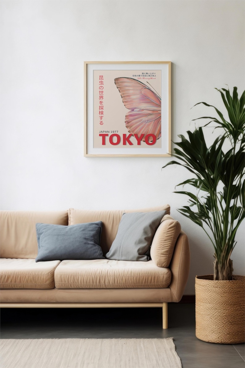 May include: A framed poster with a pink butterfly wing design and the text "JAPAN 1977 TOKYO" in red. The poster is hanging on a white wall above a beige couch with two gray pillows.