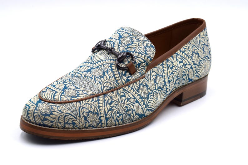 Pode incluir: Um par de sapatos de mocassim com padr&atilde;o azul e dourado, sola de couro marrom e fivela de metal prateado.
