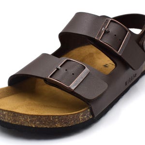 May include: Brown leather sandal with adjustable straps and buckle closures. The sandal features a cork footbed and a black sole. The brand name "mikafa" is visible on the side of the sole. This footwear is suitable for casual wear.