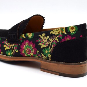 May include: Black suede loafers with a floral pattern on the front and a black suede back. The floral pattern features pink, yellow, and green flowers.