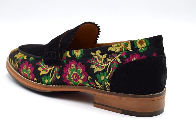 May include: Black suede loafers with a floral pattern on the front and a black suede back. The floral pattern features pink, yellow, and green flowers.