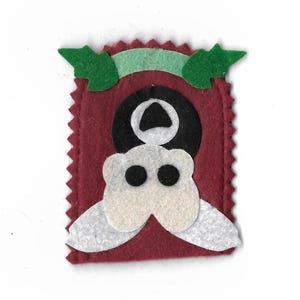 May include: A handmade felt ornament featuring a whimsical design. The ornament has a burgundy background with a black and white circular shape and a white, stylized face with black eyes. Green leaves and a light green banner are at the top.