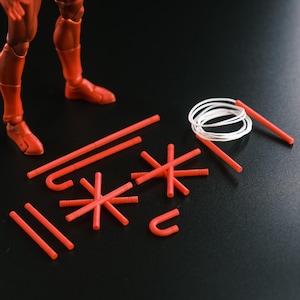 May include: A red action figure with accessories, including red rods, star-shaped pieces, and a white coiled rope. The figure's legs and feet are visible, with the accessories on a black surface.
