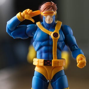 May include: A blue and yellow action figure with brown hair, wearing a visor over the eyes. The figure features a yellow harness with a circular emblem on the chest, a yellow utility belt with a red X symbol, and yellow gloves and boots.