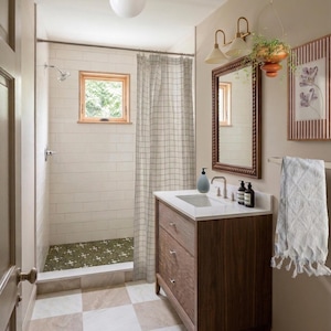 May include: A bathroom with a shower, vanity, and mirror. The shower has a checkered curtain and white tile. The vanity is dark wood with a white countertop. A framed mirror hangs above the vanity, and a towel rack is on the wall.