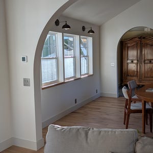 May include: Interior shot of a room with arched doorways and windows. The room has light-coloured walls, hardwood floors, and a neutral-toned sofa. Three black sconces are mounted above the windows. A wooden cabinet and round table with chairs are visible in the background.