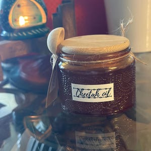 Può includere: Un barattolo di vetro marrone con un coperchio in bambù e un'etichetta bianca che dice "Cheetah oil". Il barattolo ha una superficie testurizzata ed è appoggiato su un tavolo di vetro.