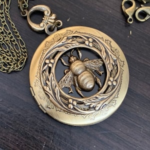 May include: A gold locket necklace featuring a bee design. The bee is encircled by a wreath of leaves and flowers. The locket is open, revealing a black interior.