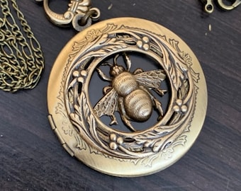 May include: A gold locket necklace with a bee design. The bee is surrounded by a wreath of leaves and flowers. The locket is open and the inside is black.
