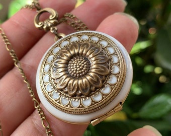 May include: A gold tone locket necklace with a white enamel sunflower design. The locket has a hinged opening.