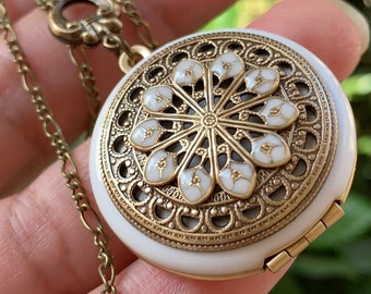 May include: A white locket necklace with a gold filigree design and a white enamel flower pattern. The locket is open and the chain is a gold curb chain.