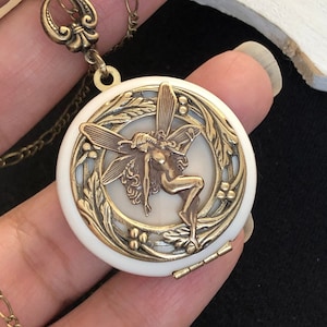 May include: A gold tone locket necklace with a white porcelain cameo featuring a fairy with wings. The locket has a hinged opening.