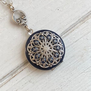 May include: A silver chain necklace with a silver locket pendant. The locket is round and has a black enamel design with a silver filigree pattern.