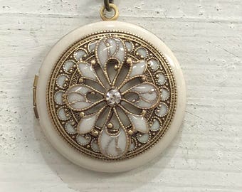 May include: A white and gold locket necklace with a floral design and a single rhinestone in the centre. The locket is round and has a delicate chain.